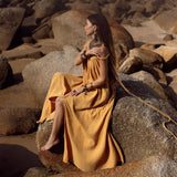 A woman wearing the Yellow Gold Greek Goddess Prom Boho Dress by AYA Sacred Wear sits on large rocks by the beach. Her long brown hair is complemented by various necklaces and bracelets, set against a natural backdrop ideal for special occasions, with ocean views and rock formations.