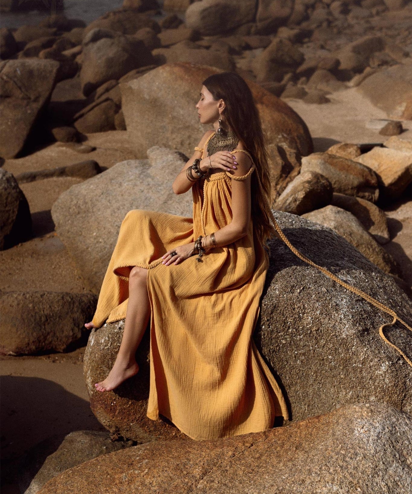 A woman wearing the Yellow Gold Greek Goddess Prom Boho Dress by AYA Sacred Wear sits on large rocks by the beach. Her long brown hair is complemented by various necklaces and bracelets, set against a natural backdrop ideal for special occasions, with ocean views and rock formations.