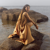 A woman in an AYA Sacred Wear Yellow Gold Greek Goddess Prom Boho Dress crouches on a rock surrounded by water, gazing serenely at the camera. The backdrop of rocks and calm water under an overcast sky enhances this special occasion in nature's embrace.