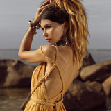A woman with long hair in a ponytail poses by the sea, wearing an AYA Sacred Wear Yellow Gold Greek Goddess Prom Boho Dress. This lightweight cotton dress with braided straps is ideal for special occasions. She holds her hair adorned with bracelets, and large rocks are visible against a cloudy sky backdrop.
