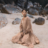 Wearing the AYA Sacred Wear Boho Dress for Women in Dusty Pink, a woman sits on a sandy beach surrounded by large rocks. Her statement earrings and bracelets complement her look as she gazes thoughtfully into the distance, her hair pulled back elegantly.