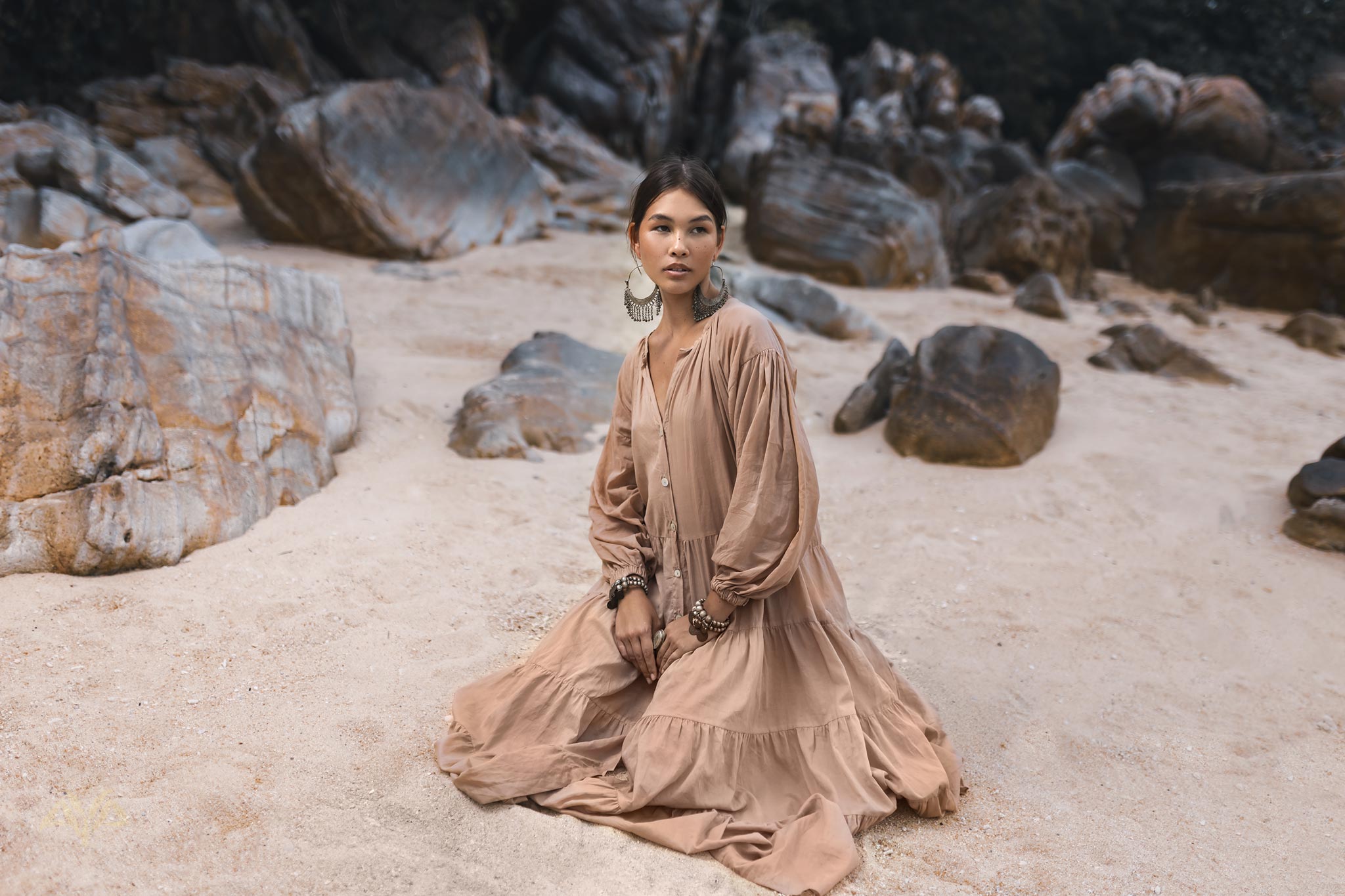 Wearing the AYA Sacred Wear Boho Dress for Women in Dusty Pink, a woman sits on a sandy beach surrounded by large rocks. Her statement earrings and bracelets complement her look as she gazes thoughtfully into the distance, her hair pulled back elegantly.