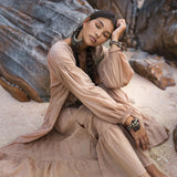 A person in a flowing AYA Sacred Wear Boho Dress for Women in dusty pink sits on sandy ground, leaning against a rock with eyes closed. They have long braided hair, large earrings, and bracelets. The rocky background adds texture to the serene setting.