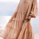 A person in a flowing dusty pink Boho Dress by AYA Sacred Wear stands by the water's edge, hand in pocket. This cotton maxi dress features buttons and layered tiers, against a serene beach backdrop with soft sunlight dancing on the water.