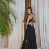 A woman stands barefoot on a light floor, facing sheer beige curtains. She looks over her shoulder, wearing the Black Oshun Dress by AYA Sacred Wear—a long, backless boho A-line made of light cotton and silk. A green palm plant is visible on the left.