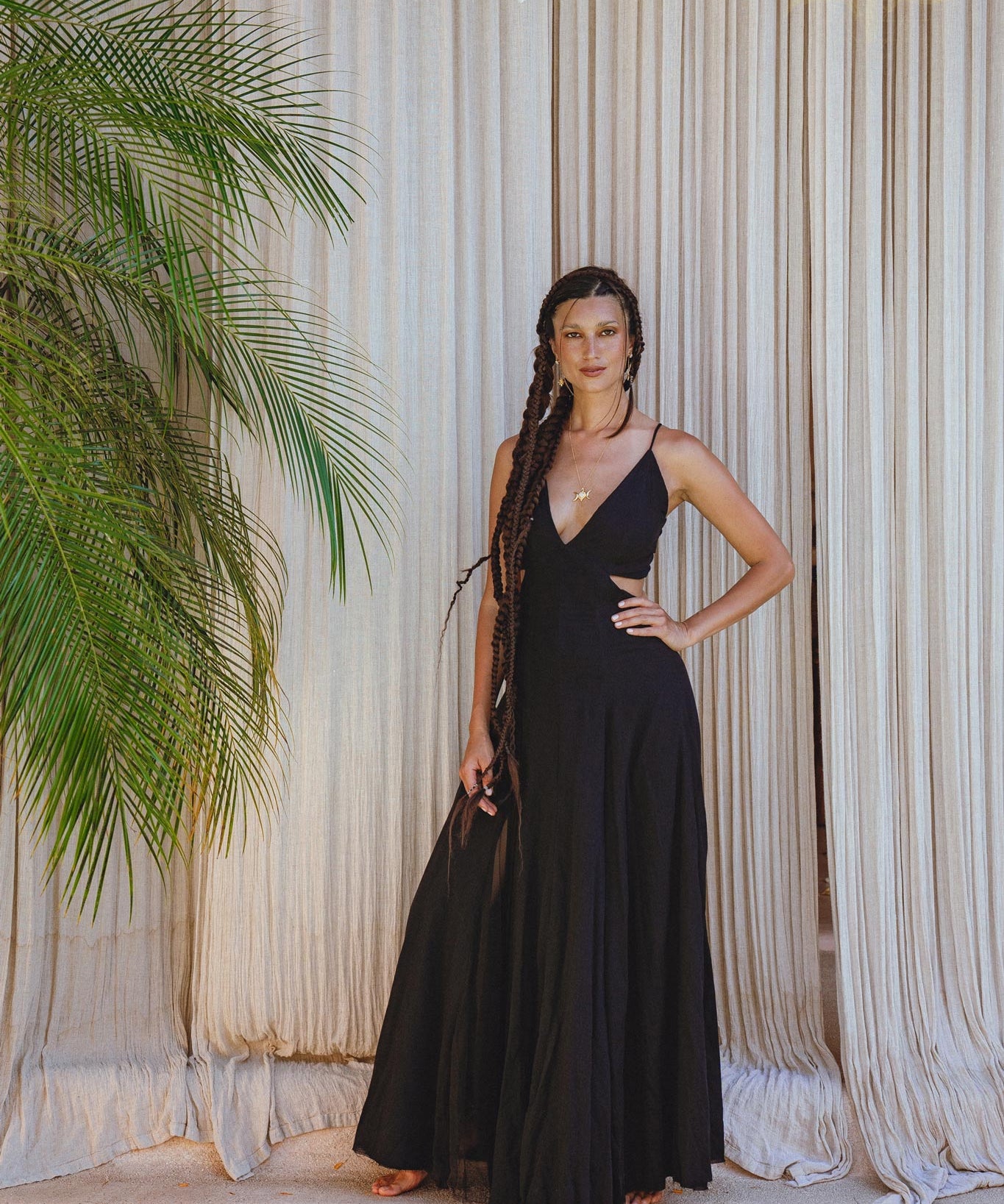 A woman poses barefoot indoors in front of beige curtains and a leafy plant, wearing the AYA Sacred Wear Black Oshun Dress—a long, black A-line formal bridal dress crafted from light cotton and silk.