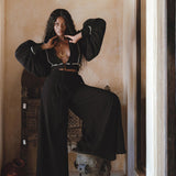 A model showcases black bohemian craft trousers from Sacred Wear, styled with a matching black cropped top. The outfit features voluminous sleeves and a relaxed fit, complemented by layered gold jewelry. The model poses confidently with one leg elevated on a decorative stool, against a warm-toned wall and elegant background elements, highlighting a casual boho aesthetic. Perfect for boho casual occasions.
