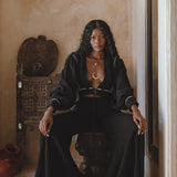 A model sitting on a decorative stool, wearing black boho pants and a matching cropped top. The outfit features flowing fabric and a relaxed fit, complemented by layered gold necklaces. The backdrop includes earthy tones and rustic decor, enhancing the bohemian aesthetic. Keywords: black boho pants, boho casual trousers, bohemian craft trousers by AYA Sacred Wear.