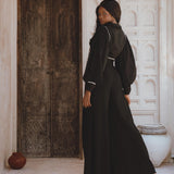 A model wearing black bohemian casual trousers from Sacred Wear stands elegantly in a rustic, white-walled interior, showcasing a flowing silhouette and paired with a matching top. The backdrop features carved wooden doors and decorative pottery, enhancing the boho aesthetic.