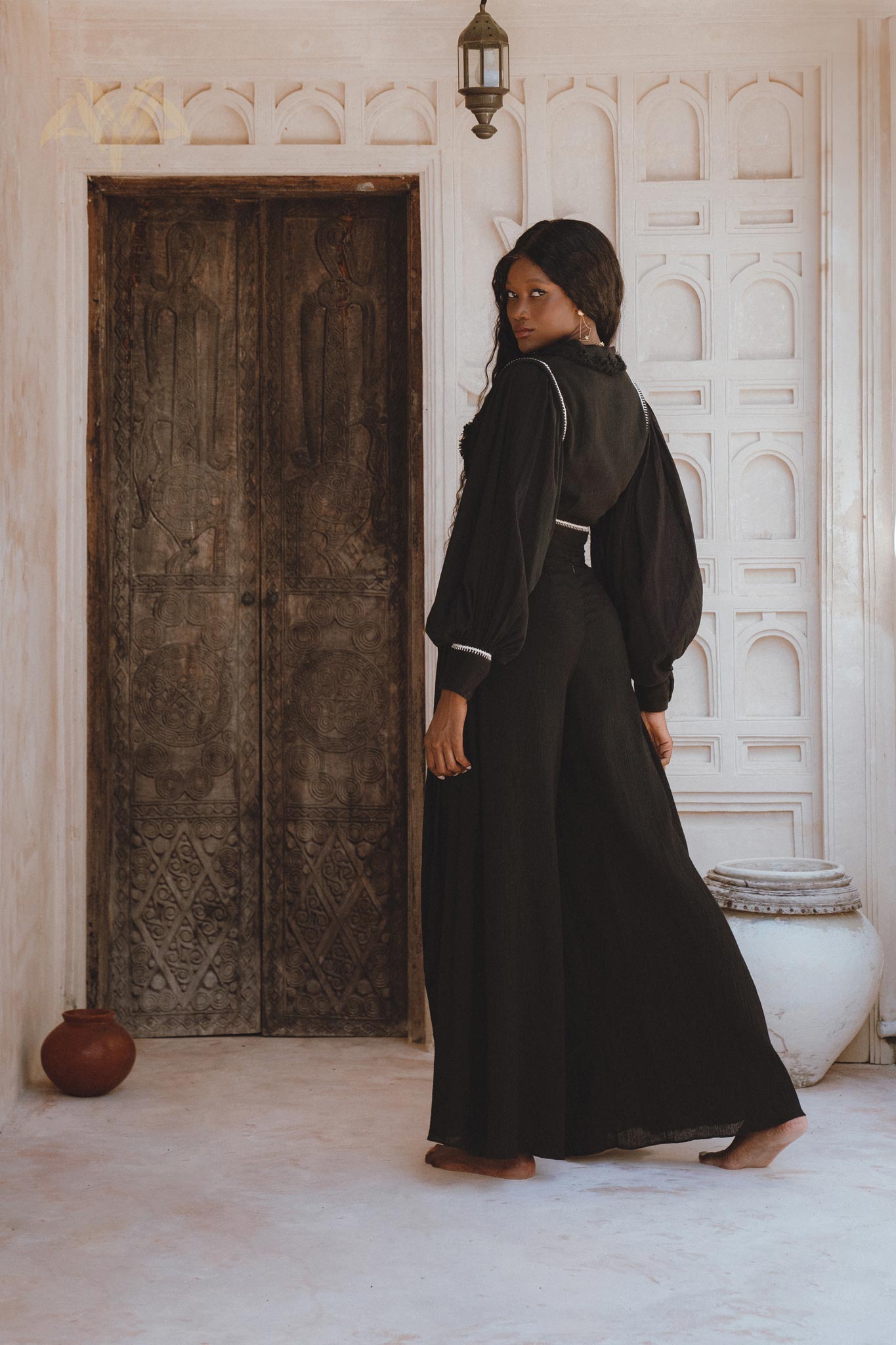 A model wearing black bohemian casual trousers from Sacred Wear stands elegantly in a rustic, white-walled interior, showcasing a flowing silhouette and paired with a matching top. The backdrop features carved wooden doors and decorative pottery, enhancing the boho aesthetic.