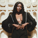 A model poses elegantly against a textured wall, wearing stylish black bohemian trousers paired with a statement blouse featuring billowing sleeves. She accessorizes with layered necklaces, showcasing a chic and casual boho look perfect for any occasion. The sunny ambiance highlights the outfit's details, emphasizing its craftsmanship and relaxed vibe. Perfect for those searching for boho casual trousers.