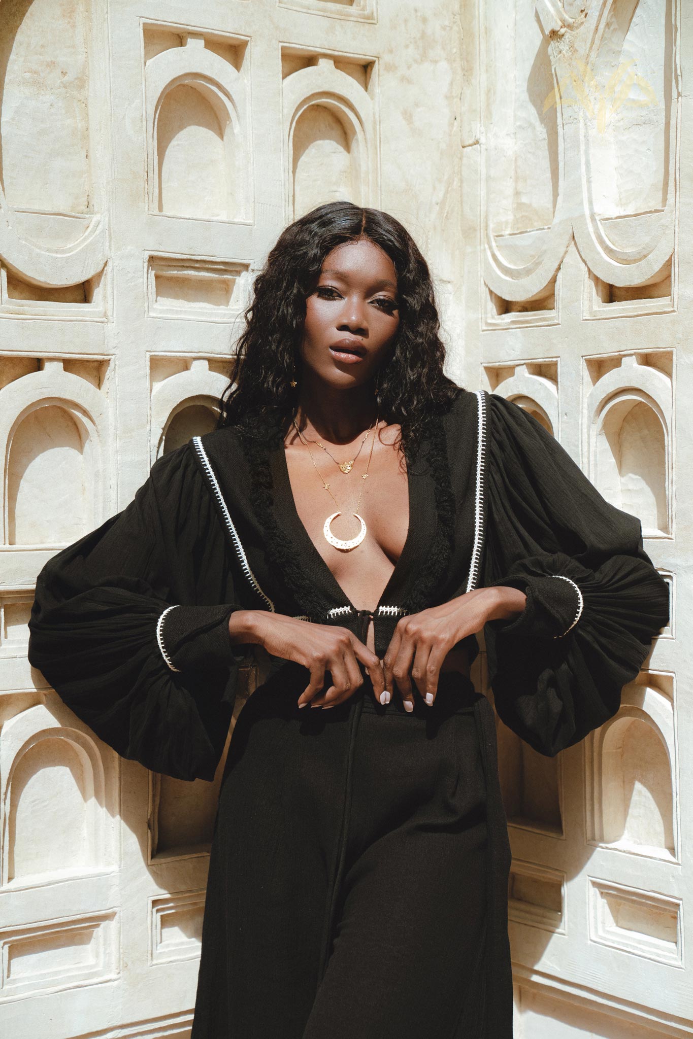 A model poses elegantly against a textured wall, wearing stylish black bohemian trousers paired with a statement blouse featuring billowing sleeves. She accessorizes with layered necklaces, showcasing a chic and casual boho look perfect for any occasion. The sunny ambiance highlights the outfit's details, emphasizing its craftsmanship and relaxed vibe. Perfect for those searching for boho casual trousers.