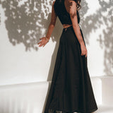 A woman in an elegant Black Prom Handmade Dress from AYA Sacred Wear, featuring a boho design with a black sleeveless top and long handwoven skirt, leans against a white wall. Dappled shadows of leaves adorn the wall and her figure as she gazes contemplatively to the side.