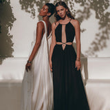 Against the textured wall outdoors, two models showcase elegant dresses made from handwoven fabrics. One model is adorned in a flowing white gown, while the other wears a stylish black prom dress with cutouts, crafted by AYA Sacred Wear and ideal for boho bridesmaids. Shadows of leaves cast a patterned backdrop on the wall.