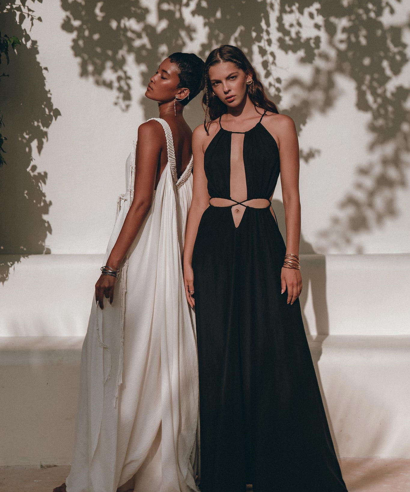 Against the textured wall outdoors, two models showcase elegant dresses made from handwoven fabrics. One model is adorned in a flowing white gown, while the other wears a stylish black prom dress with cutouts, crafted by AYA Sacred Wear and ideal for boho bridesmaids. Shadows of leaves cast a patterned backdrop on the wall.