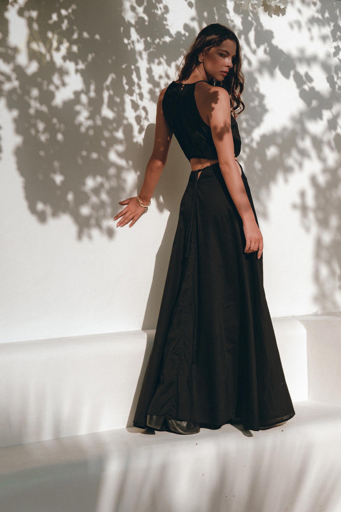 A woman in an elegant Black Prom Handmade Dress from AYA Sacred Wear, featuring a boho design with a black sleeveless top and long handwoven skirt, leans against a white wall. Dappled shadows of leaves adorn the wall and her figure as she gazes contemplatively to the side.