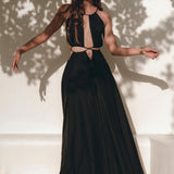 A woman exudes confidence as she stands in the sunlight wearing the Black Prom Handmade Dress by AYA Sacred Wear, a long, sleeveless black dress featuring cut-out details. The shadows of leaves play on the wall behind her. Her long, wavy hair complements the organic fabric bracelets on her left wrist, showcasing both elegance and sustainability.