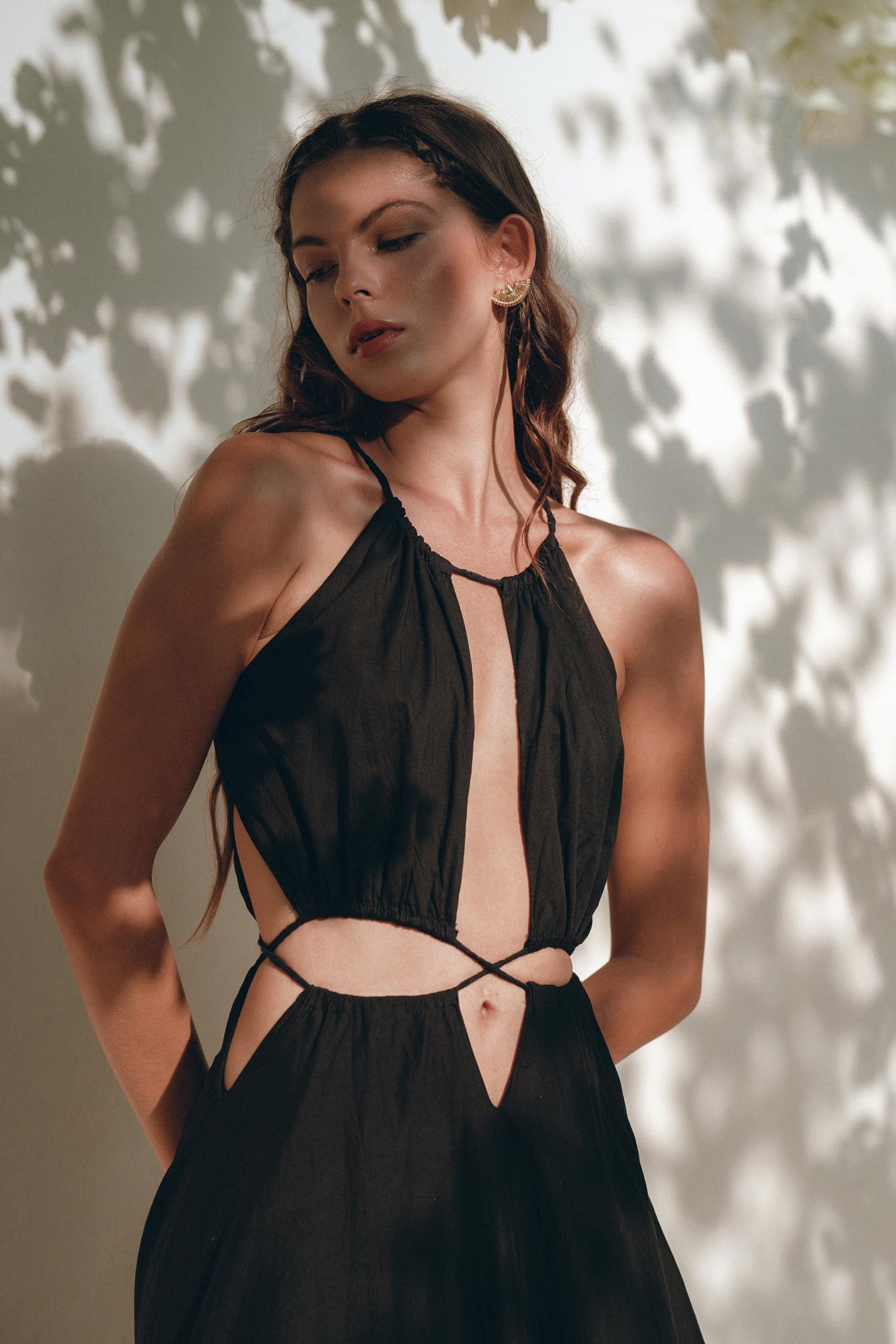A woman showcases the Black Prom Handmade Dress from AYA Sacred Wear, a boho dress with cut-out details crafted from organic fabric. She stands against a wall playfully illuminated by sunlight and shadow, gazing downward, with her long wavy hair cascading over her shoulder.