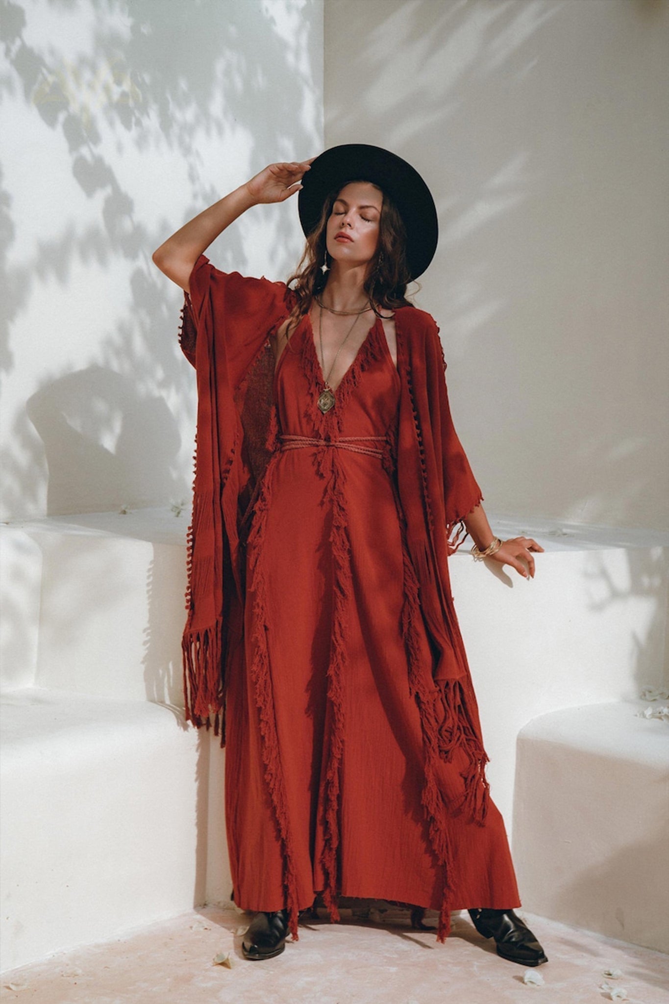 Full-length fashion shot of a model wearing the divine Wine Red raw cotton dress layered with a matching fringe poncho and a black wide-brimmed hat, leaning against a white architectural wall.
