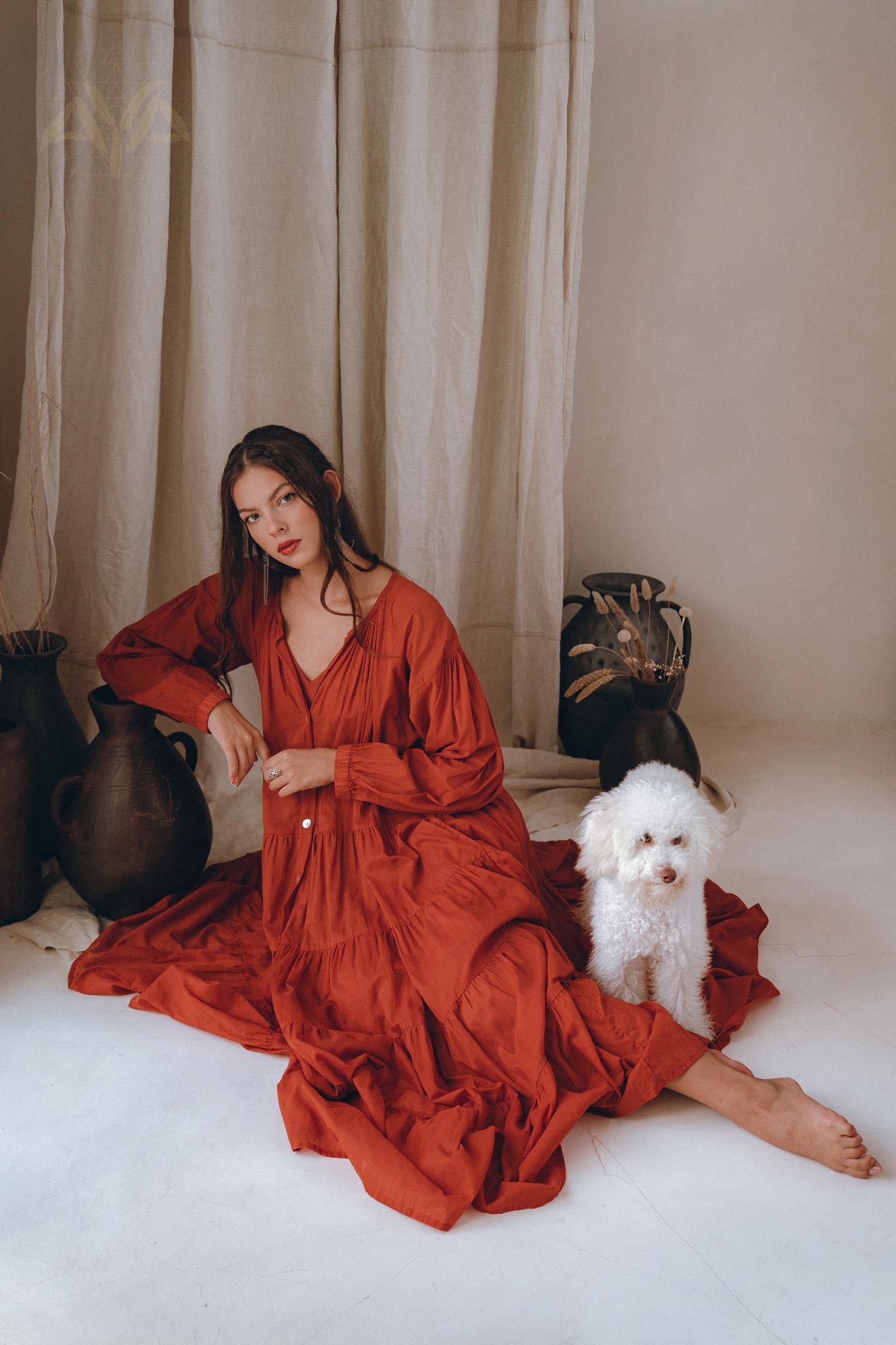 A woman wearing an AYA Sacred Wear Wine Red Boho Prom Dress gracefully sits on the floor against a beige curtain backdrop. Next to her is a small, fluffy white dog, with dark decorative vases arranged nearby.