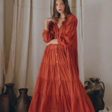 A woman wearing a Boho Dress for Women by AYA Sacred Wear, specifically the Wine Red Boho Prom Dress, stands barefoot in front of beige curtains. She gracefully holds part of her cotton Light Summer Dress with one hand, as ceramic pots rest on the floor beside her. Her long hair flows freely.
