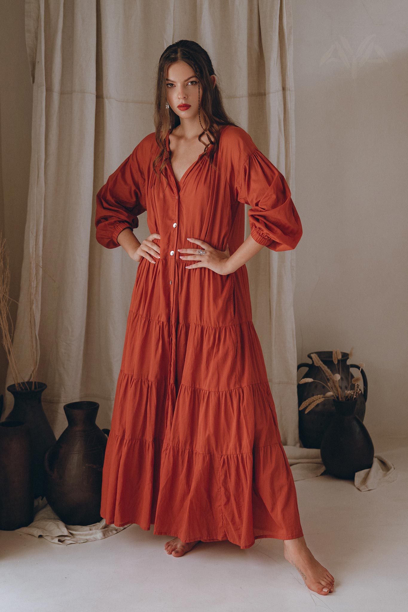 A woman stands confidently in a flowing Wine Red Boho Prom Dress with long sleeves, from AYA Sacred Wear. Her hands are on her hips, exuding poise. The background features neutral-toned curtains and decorative vases, enhancing her stylish presence in this Boho Dress for Women.