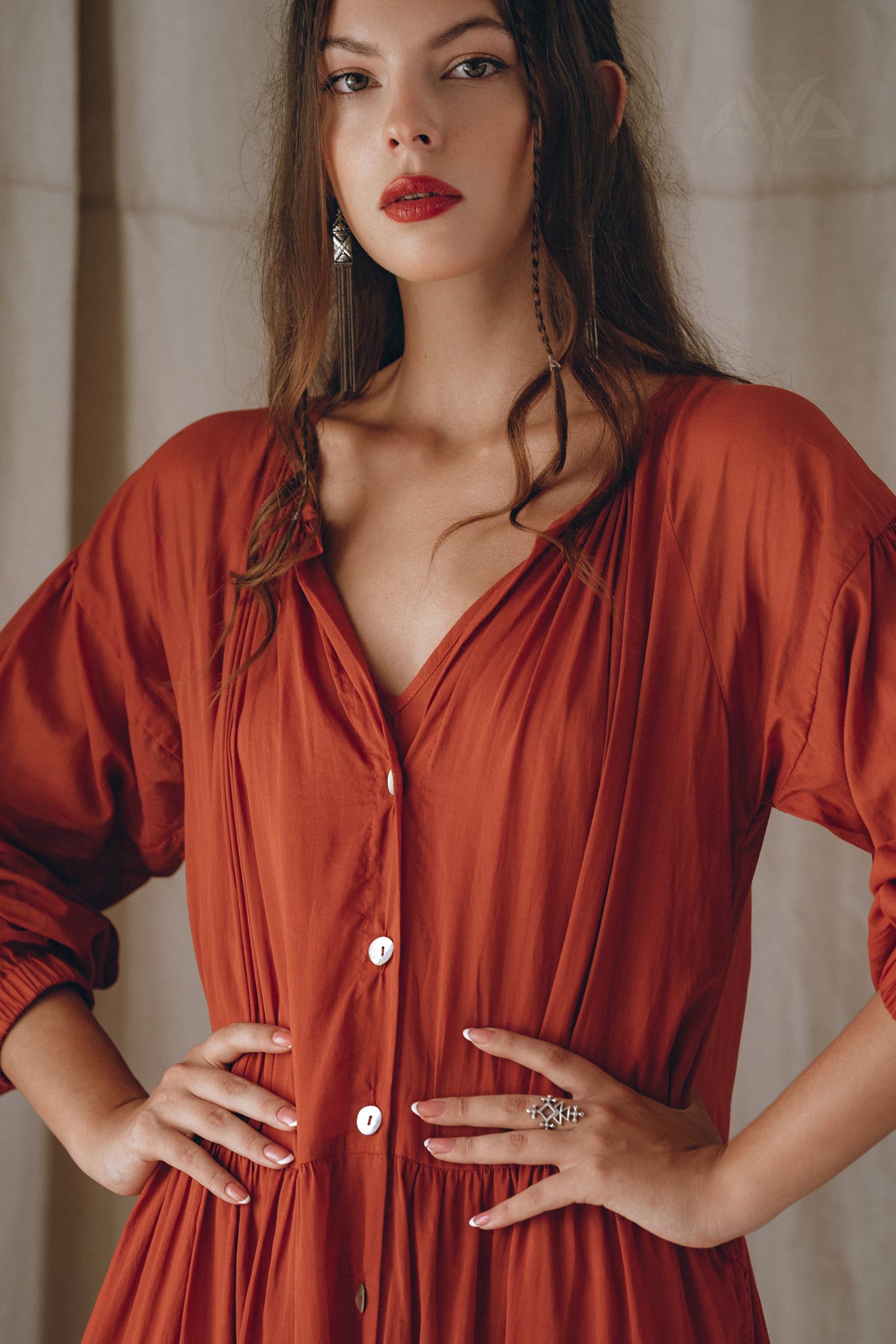 A woman with long brown hair and red lipstick stands confidently with her hands on her hips. She is wearing an AYA Sacred Wear Boho Dress for Women in a flowing wine red color, adorned with buttons and crafted from elegant cotton fabric. Complementing her look are silver earrings, while the background features softly draped fabric.
