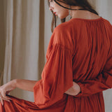 A woman with long hair is turned away from the camera, wearing the Boho Dress for Women by AYA Sacred Wear. The flowing, wine red dress features puffy sleeves and is made of cotton. She elegantly holds out the fabric in a scene bathed in soft natural lighting.
