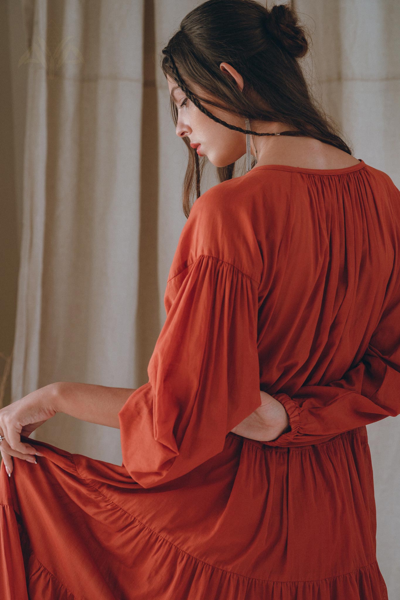 A woman with long hair is turned away from the camera, wearing the Boho Dress for Women by AYA Sacred Wear. The flowing, wine red dress features puffy sleeves and is made of cotton. She elegantly holds out the fabric in a scene bathed in soft natural lighting.