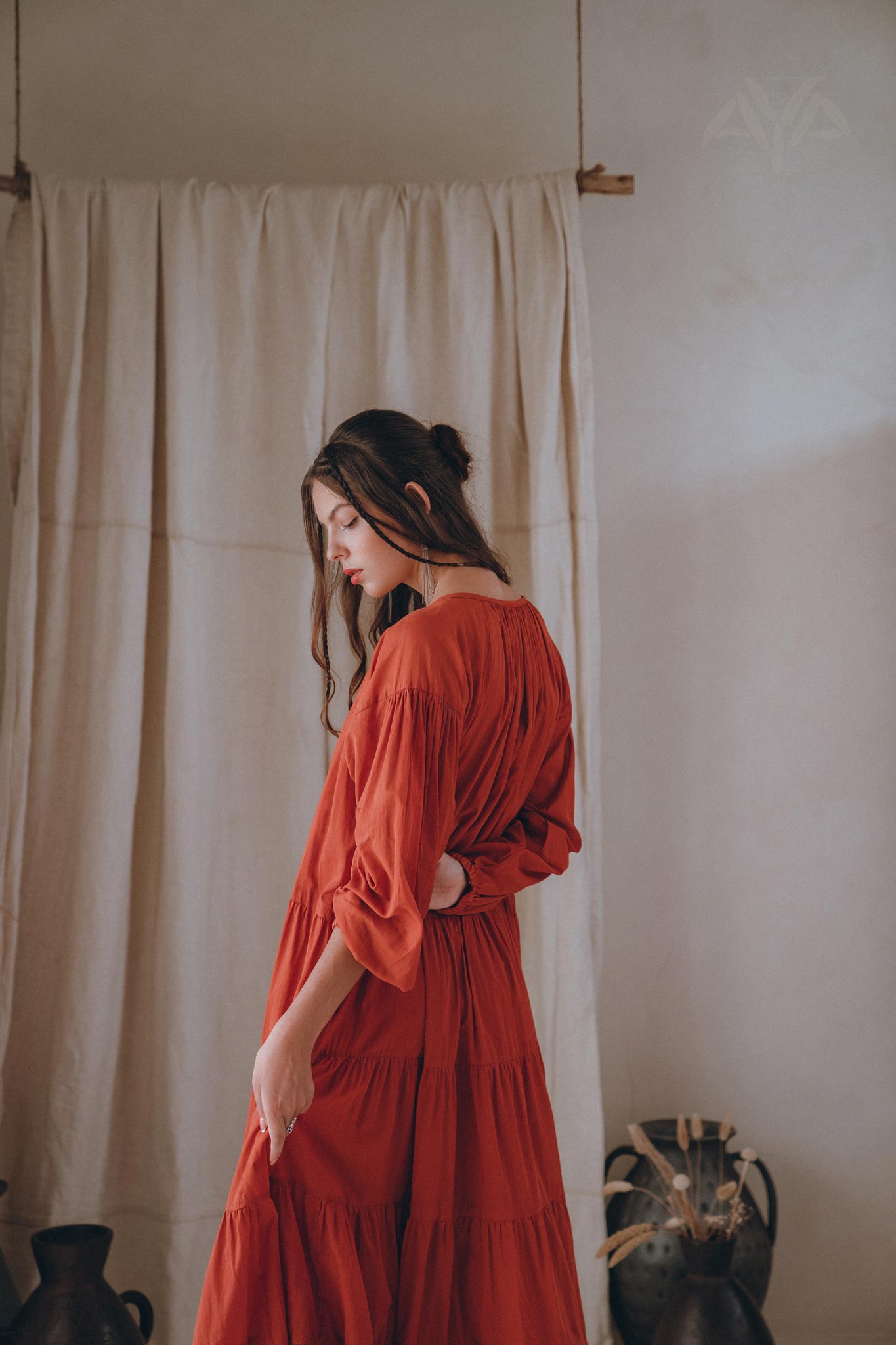 A woman wearing the AYA Sacred Wear Wine Red Boho Prom Dress stands sideways in a softly lit room. Her hair is tied back as she gazes downward, surrounded by a neutral-toned curtain and pottery with dried plants.