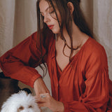 A woman wearing the AYA Sacred Wear's Wine Red Boho Prom Dress gently interacts with a small white fluffy dog. She is sitting indoors against a light-colored curtain backdrop, looking thoughtful with her hair styled in loose waves. The soft cotton fabric of the Boho Dress for Women adds to the serene ambiance of the scene.