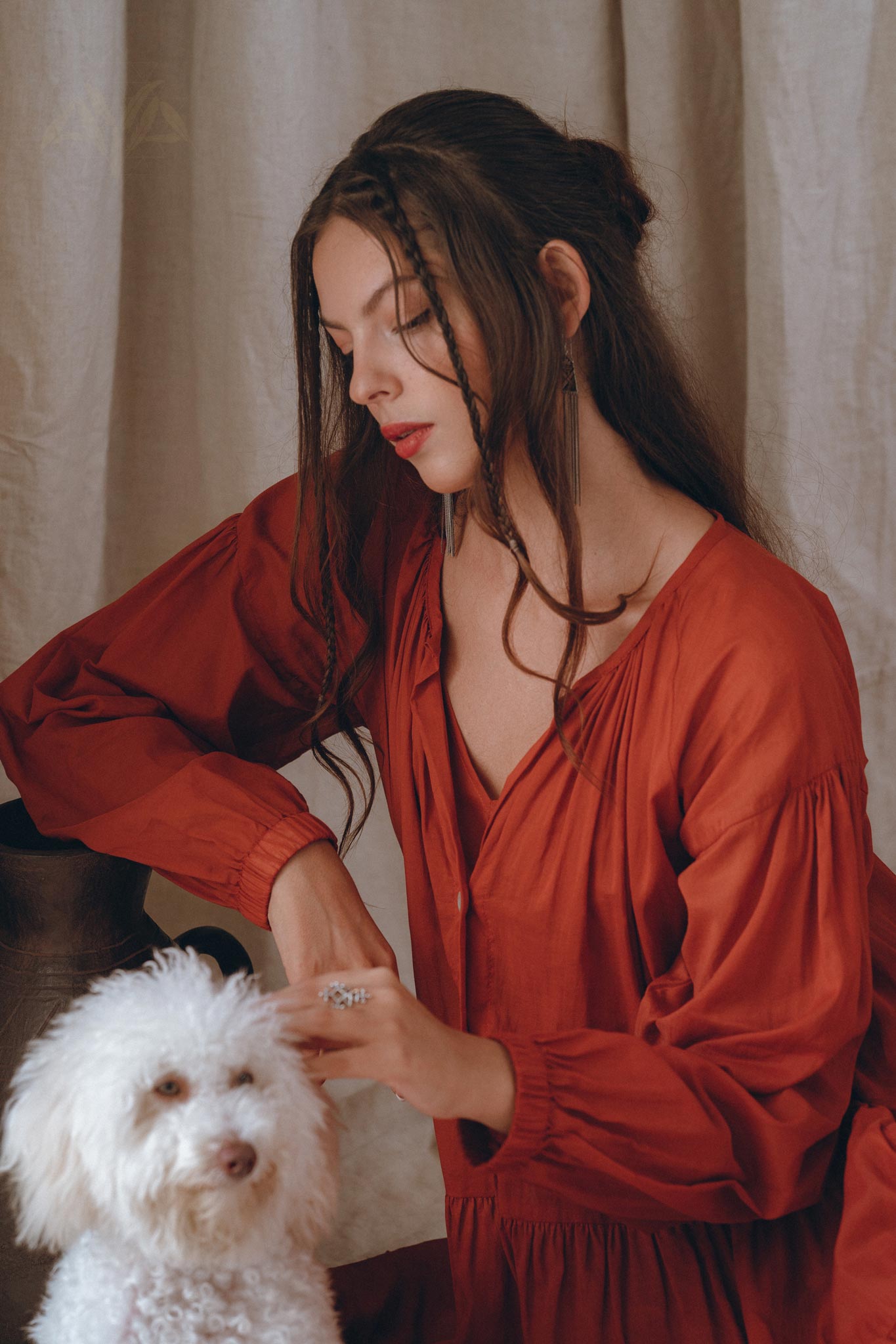 A woman wearing the AYA Sacred Wear's Wine Red Boho Prom Dress gently interacts with a small white fluffy dog. She is sitting indoors against a light-colored curtain backdrop, looking thoughtful with her hair styled in loose waves. The soft cotton fabric of the Boho Dress for Women adds to the serene ambiance of the scene.