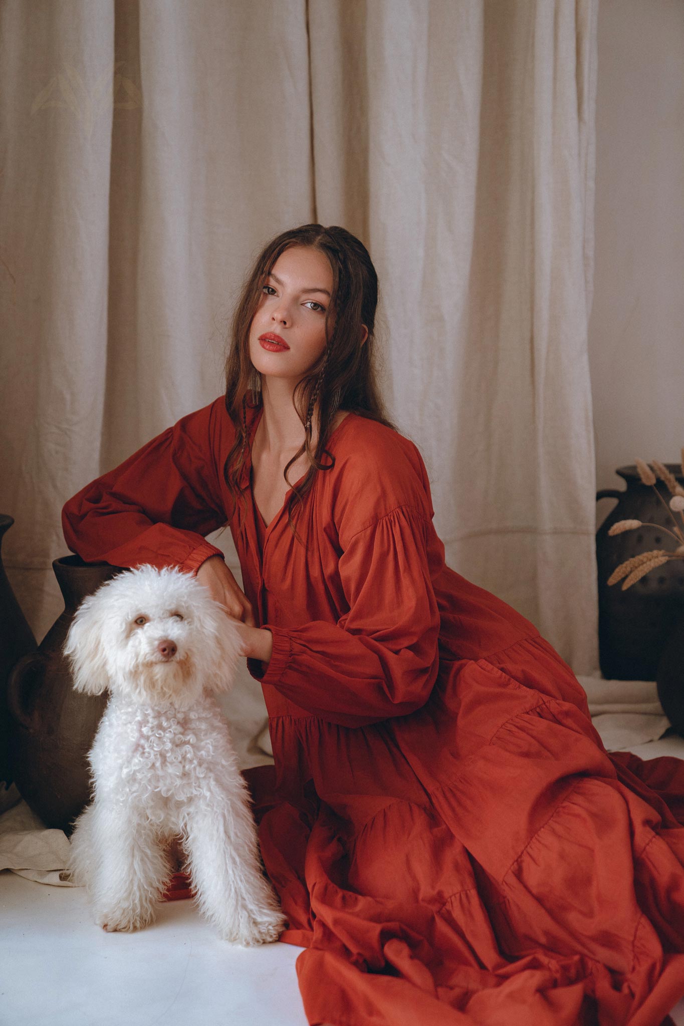 A woman in a beautiful Wine Red Boho Prom Dress from AYA Sacred Wear sits on the floor next to a fluffy white dog. The background features neutral-colored curtains and decorative pottery, creating a warm, elegant atmosphere.