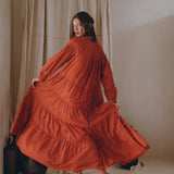 A woman elegantly poses in a wine red Boho Dress for Women by AYA Sacred Wear, her flowing maxi dress gracefully cascading as she slightly raises one foot. She stands barefoot on a light floor against a neutral curtain backdrop, creating a soft and sophisticated atmosphere.