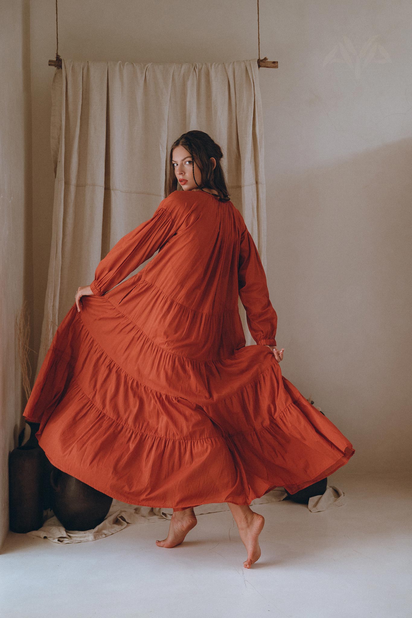 A woman elegantly poses in a wine red Boho Dress for Women by AYA Sacred Wear, her flowing maxi dress gracefully cascading as she slightly raises one foot. She stands barefoot on a light floor against a neutral curtain backdrop, creating a soft and sophisticated atmosphere.