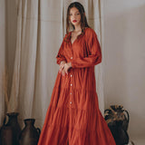 A person wearing the AYA Sacred Wear Wine Red Boho Prom Dress, a flowing light summer dress, stands barefoot in front of cream-colored drapes. The setting includes pottery and dried plants, creating a rustic, serene atmosphere.