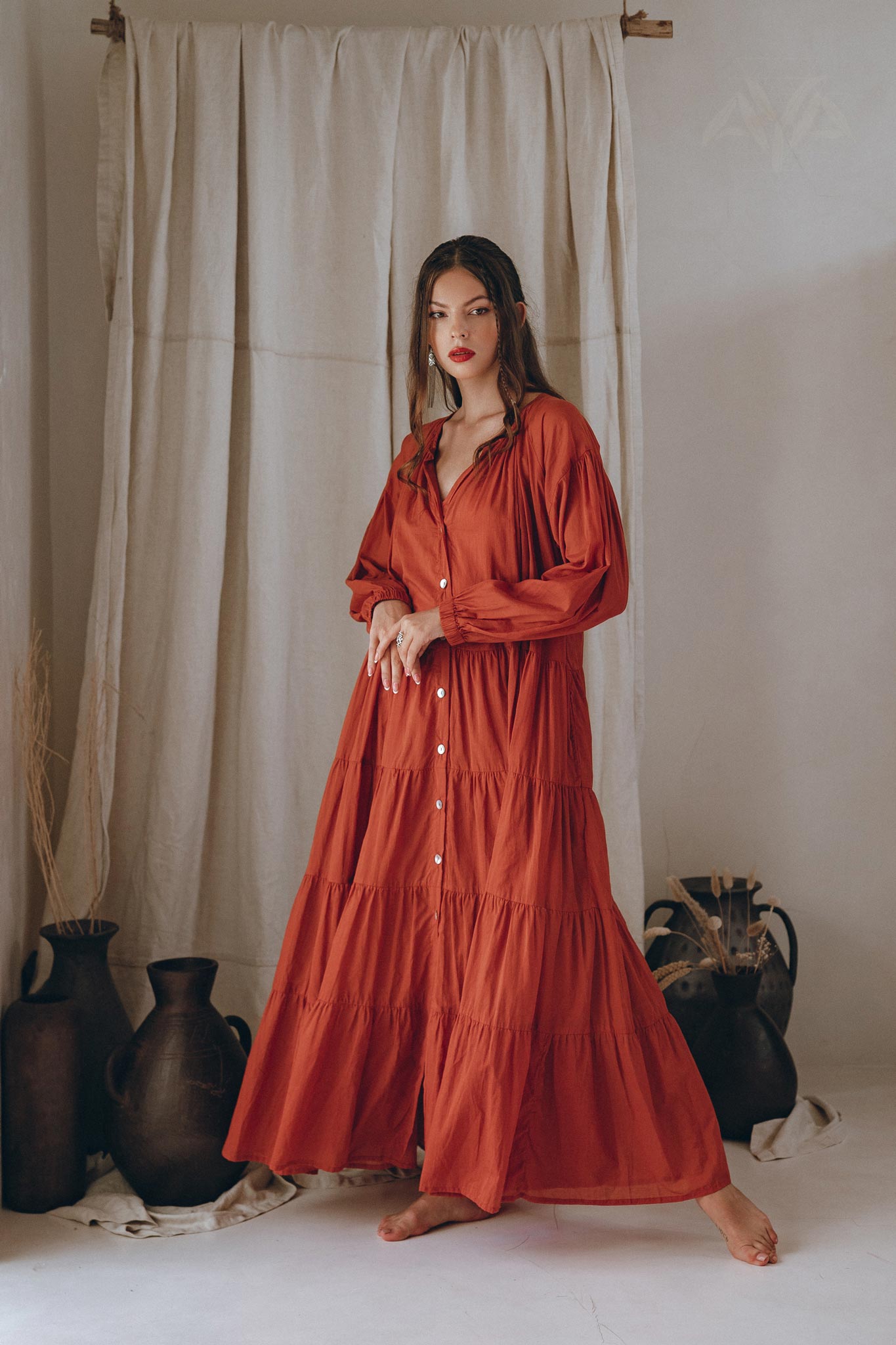 A person wearing the AYA Sacred Wear Wine Red Boho Prom Dress, a flowing light summer dress, stands barefoot in front of cream-colored drapes. The setting includes pottery and dried plants, creating a rustic, serene atmosphere.