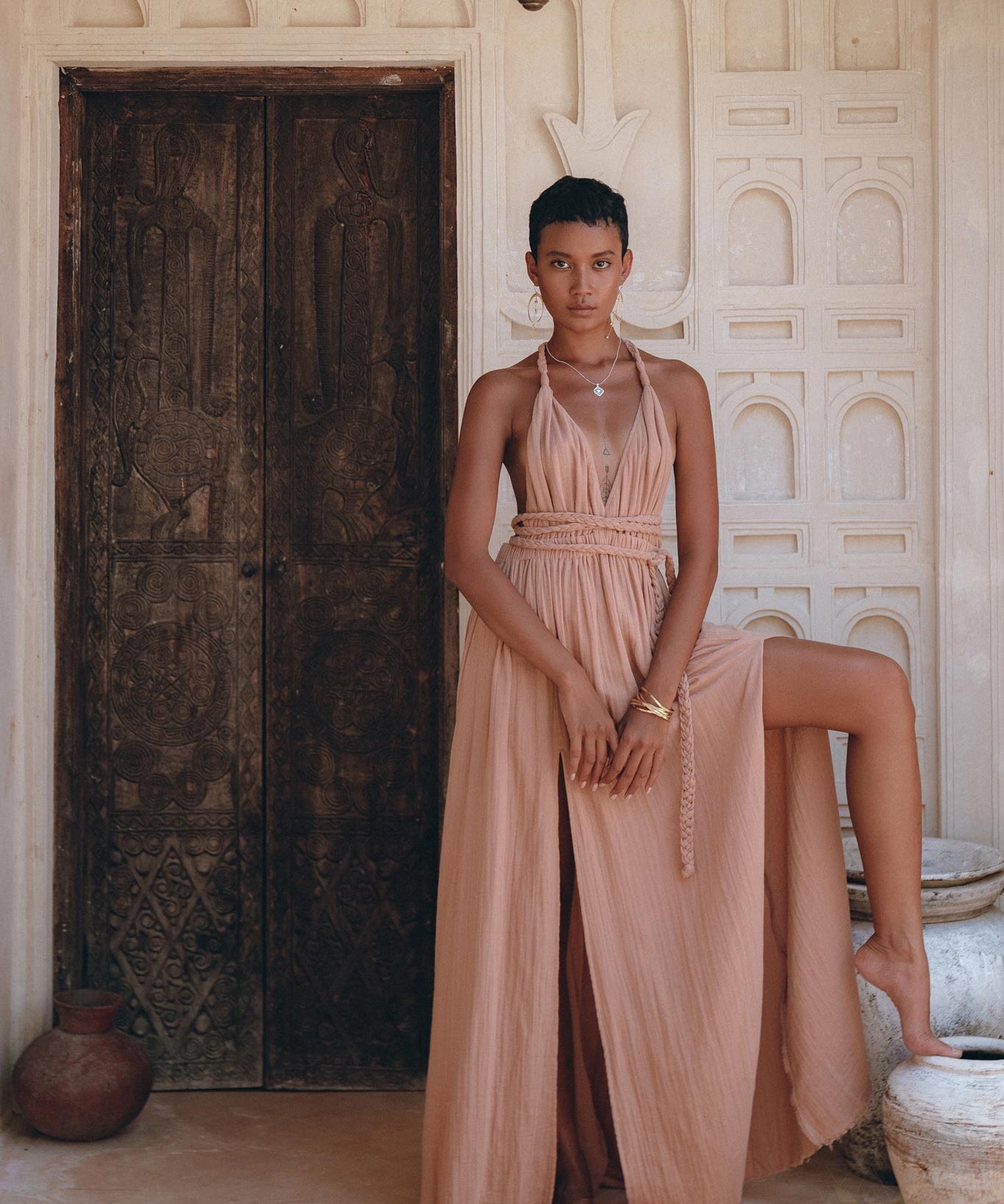 A person wearing the Dusty Pink Greek Goddess Dress by AYA Sacred Wear stands in front of an intricately carved wooden door. The background features ornate white plasterwork. With short hair and one leg raised, they elegantly showcase the dress's high slit.