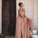 Dressed in a Dusty Pink Greek Goddess Dress from AYA Sacred Wear, a person poses elegantly against an ornate wooden door adorned with intricate carvings. They rest one foot on a clay pot and gaze thoughtfully to the side. The setting is beautifully accented with terracotta pots and Arabic-style architecture.