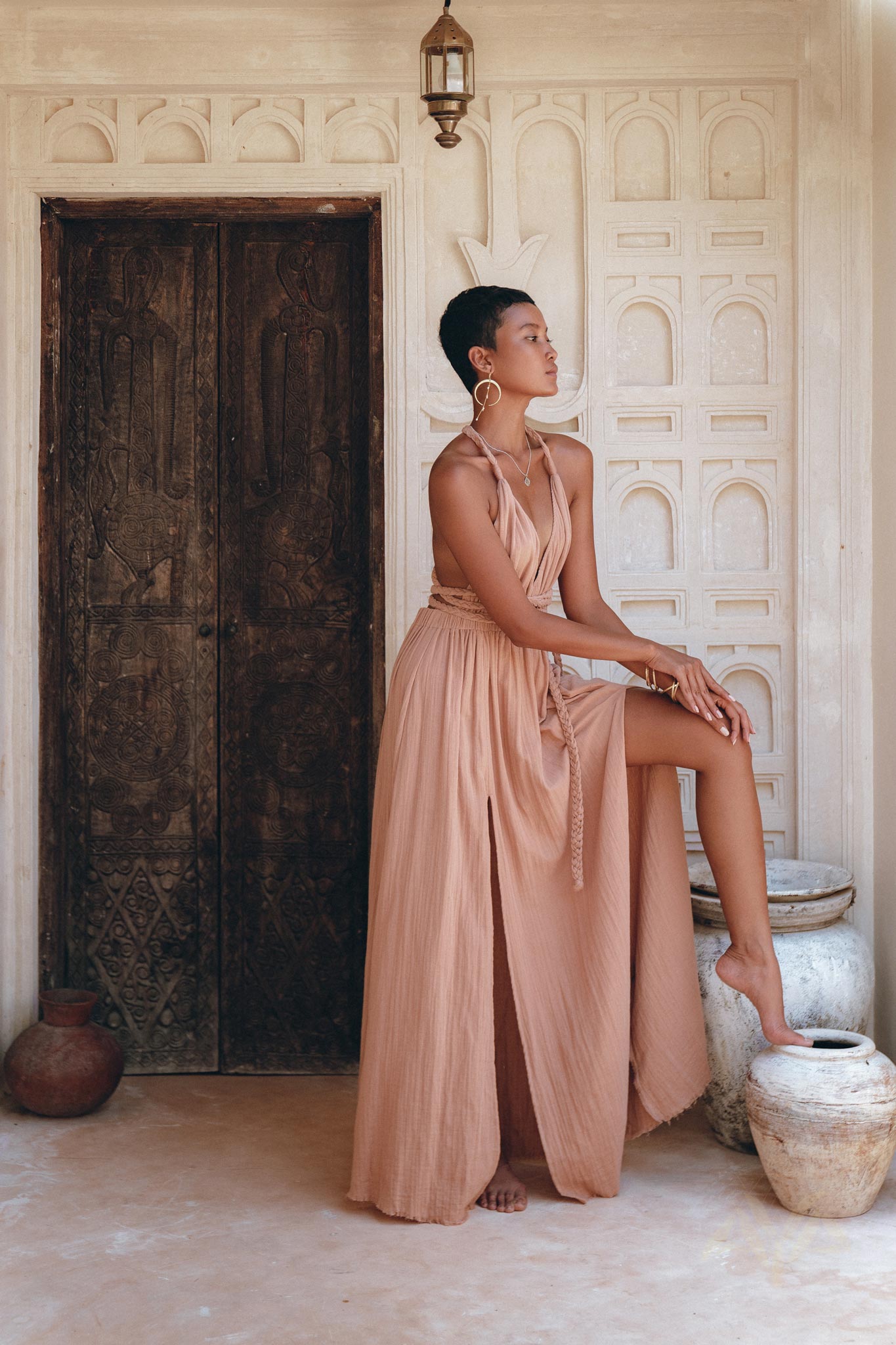 Dressed in a Dusty Pink Greek Goddess Dress from AYA Sacred Wear, a person poses elegantly against an ornate wooden door adorned with intricate carvings. They rest one foot on a clay pot and gaze thoughtfully to the side. The setting is beautifully accented with terracotta pots and Arabic-style architecture.