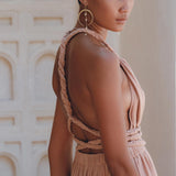A person with short hair wearing the Dusty Pink Greek Goddess Dress by AYA Sacred Wear, styled with large hoop earrings, stands in a softly lit room looking away. The background showcases an ornate, light-colored wall adorned with arch patterns.