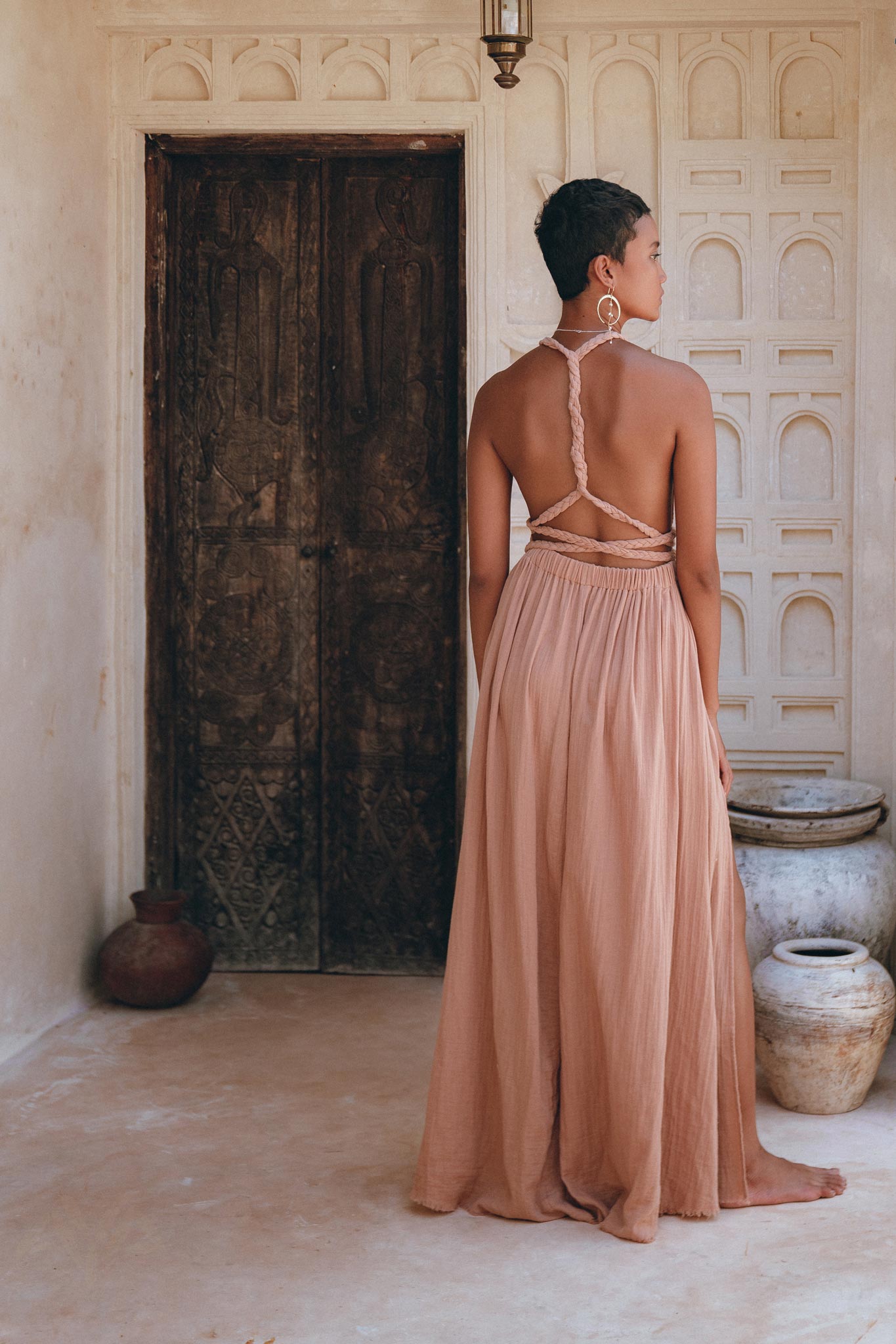 A person stands with their back to the camera, adorned in a Dusty Pink Greek Goddess Dress by AYA Sacred Wear, featuring braided straps and a long, flowing design. They are positioned before a rustic wooden door, encircled by textured beige walls and decorative pots.