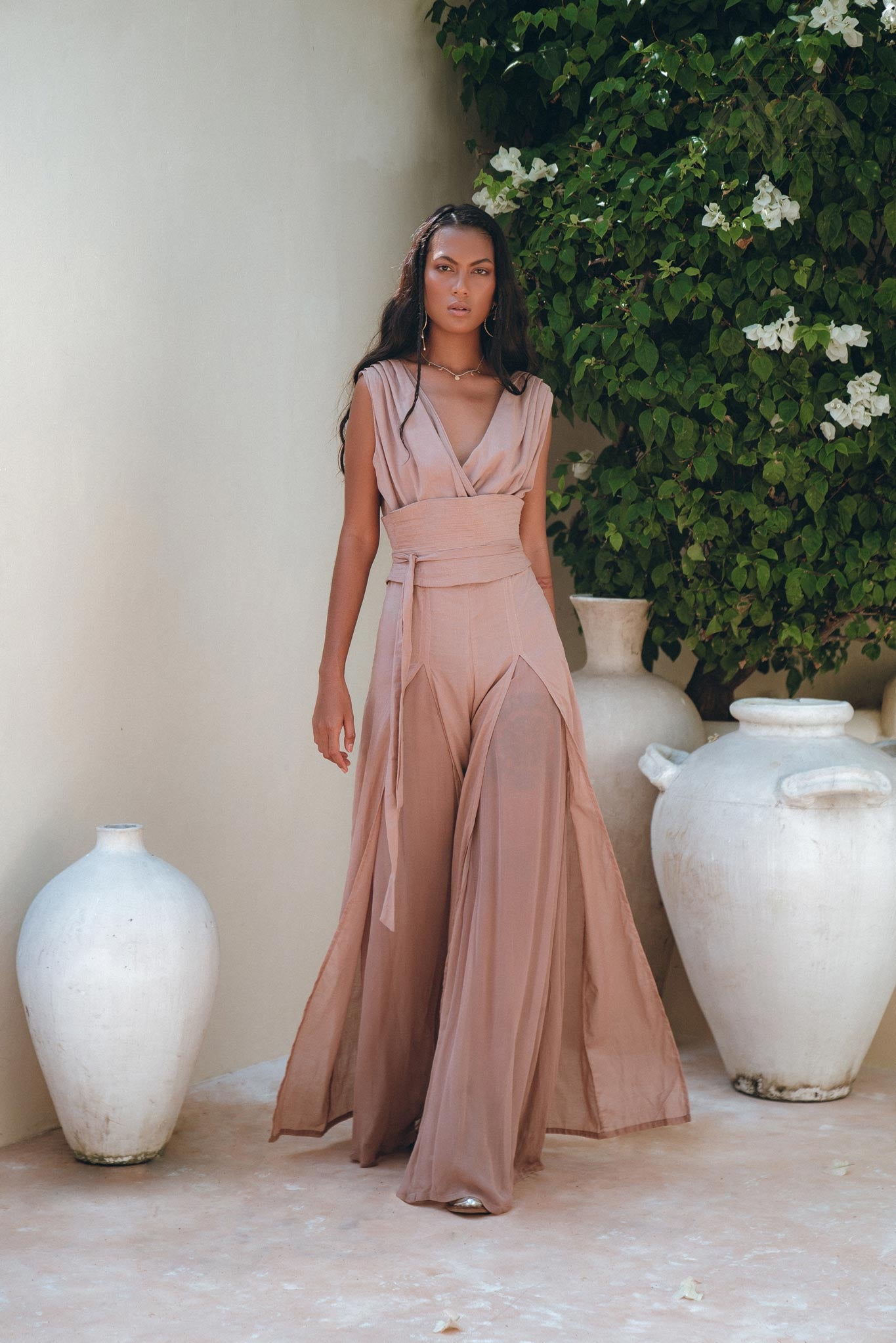 Female model walking elegantly in a dusty pink organic cotton and silk wide-leg jumpsuit, surrounded by white rustic vases.