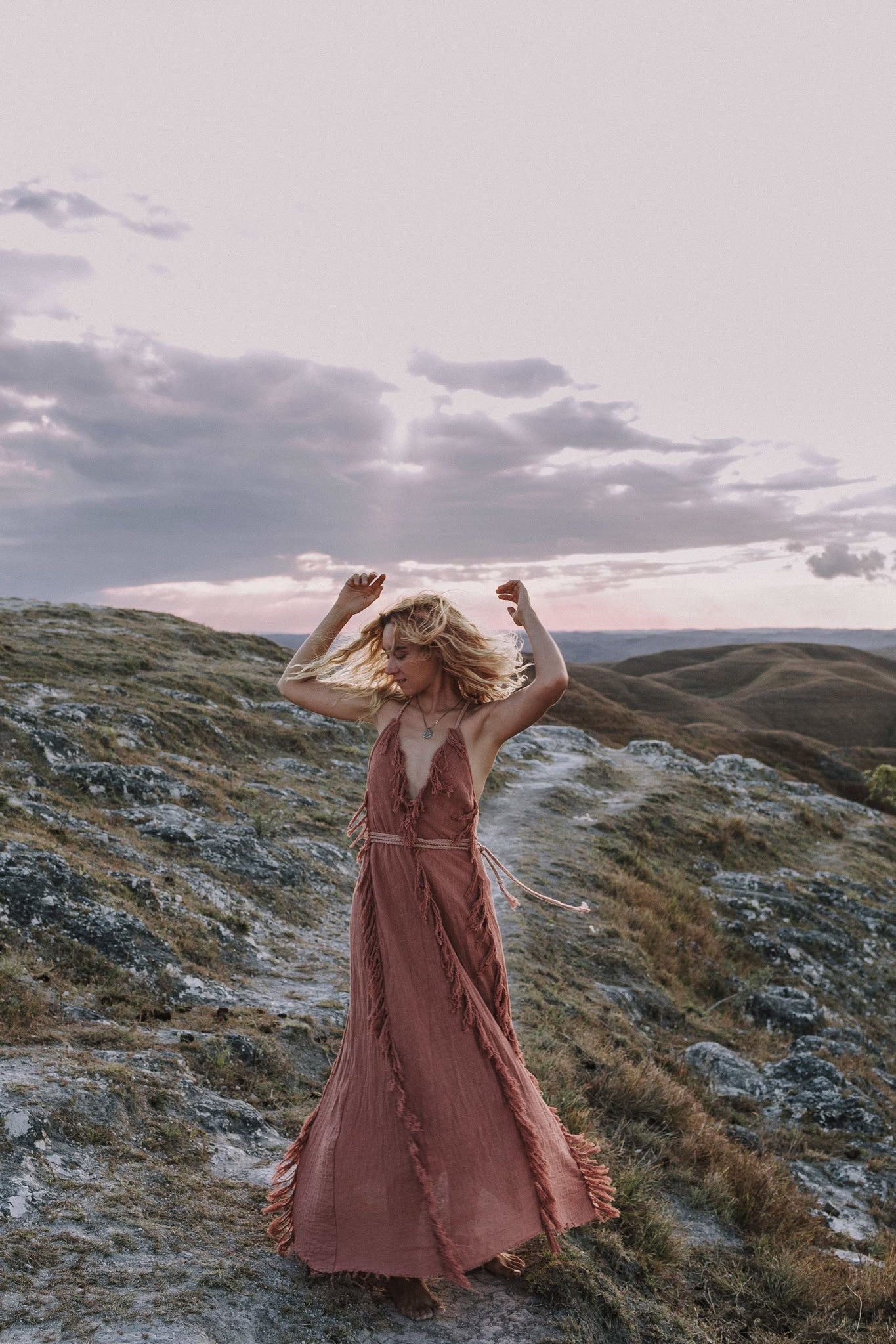 A model wearing the delicate Dusty Rose raw cotton goddess dress by AYA Sacred Wear, standing on a rocky hill against a beautiful twilight sky.