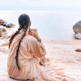 A person with long braided hair sits on a sandy beach, wearing an AYA Sacred Wear Boho Dress for Women in Dusty Pink, as they gaze at the calm sea under a cloudy sky. Large rocks are visible in the background.