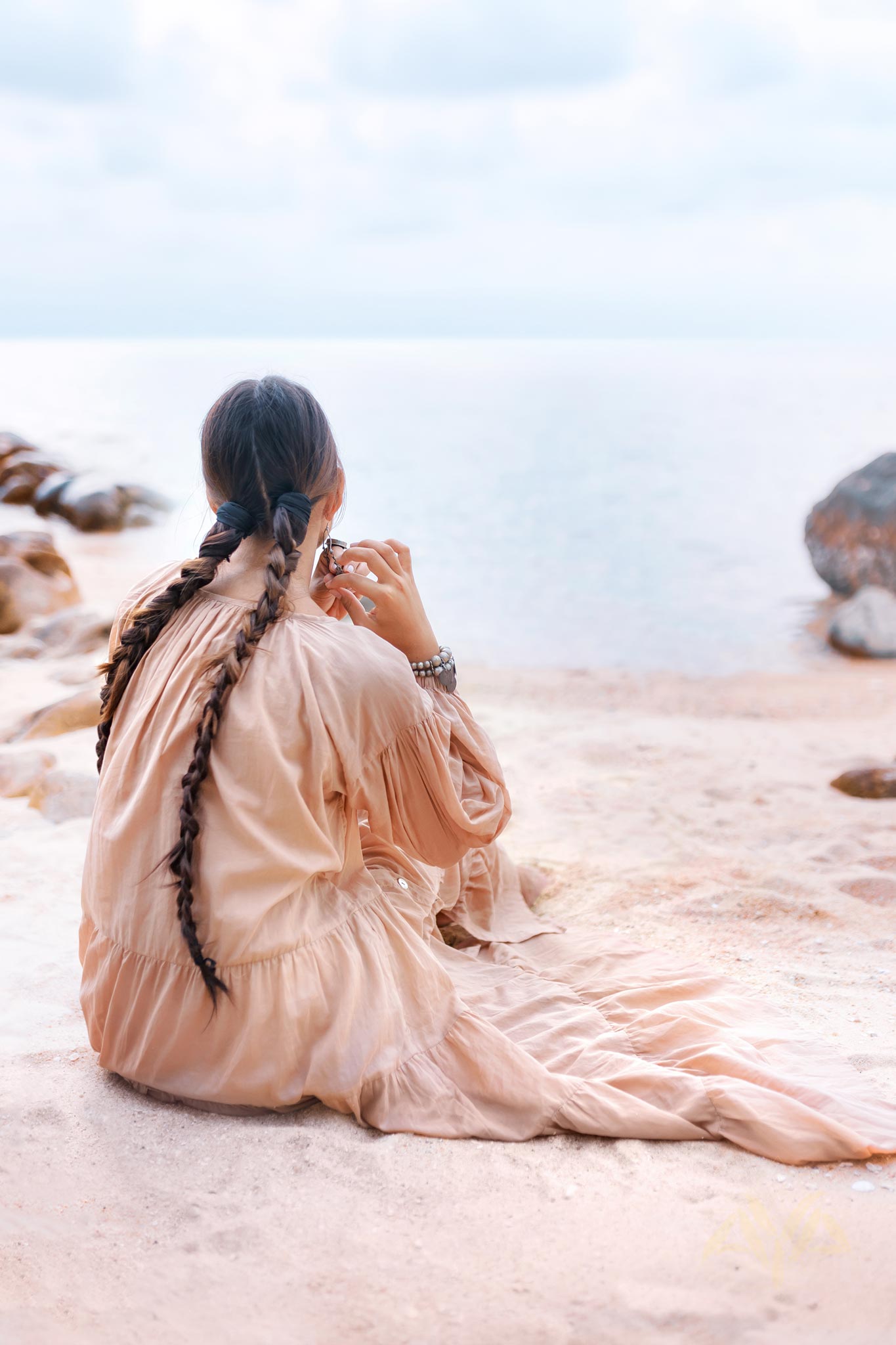 A person with long braided hair sits on a sandy beach, wearing an AYA Sacred Wear Boho Dress for Women in Dusty Pink, as they gaze at the calm sea under a cloudy sky. Large rocks are visible in the background.