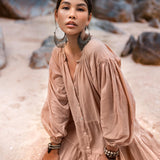 A woman kneels on a sandy beach in an AYA Sacred Wear Boho Dress, Dusty Pink. Adorned with large hoop earrings and bracelets, her light summer dress of soft cotton complements the serene rocky backdrop as she gazes at the camera.