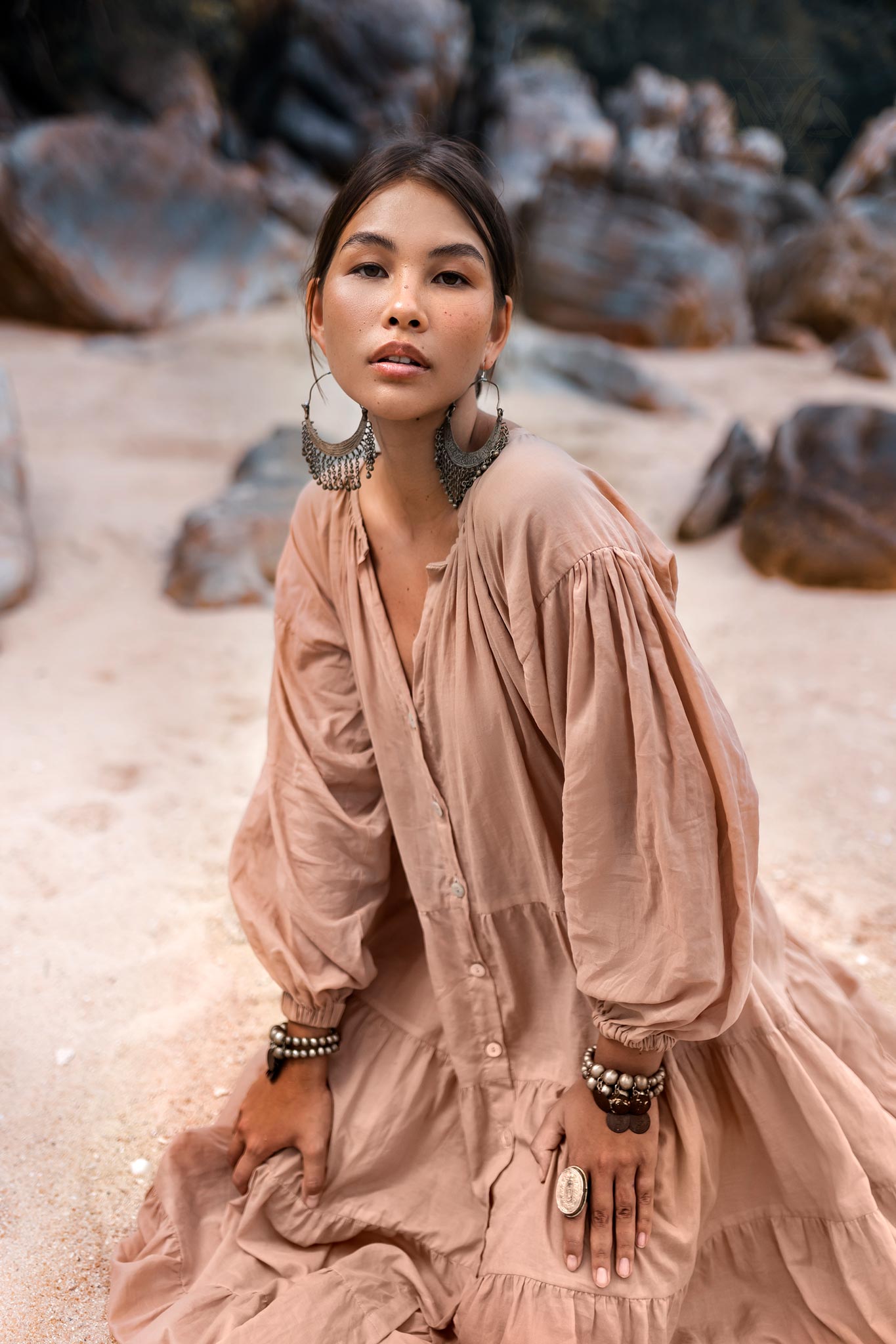 A woman kneels on a sandy beach in an AYA Sacred Wear Boho Dress, Dusty Pink. Adorned with large hoop earrings and bracelets, her light summer dress of soft cotton complements the serene rocky backdrop as she gazes at the camera.