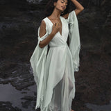 Front view of a model walking in a lush tropical garden wearing the ice blue Makosh goddess dress. Features flowing silk wing sleeves and textured organic crinkle cotton wrap. High-quality sustainable wedding gown for a celestial bridal look by AYA Sacred Wear.