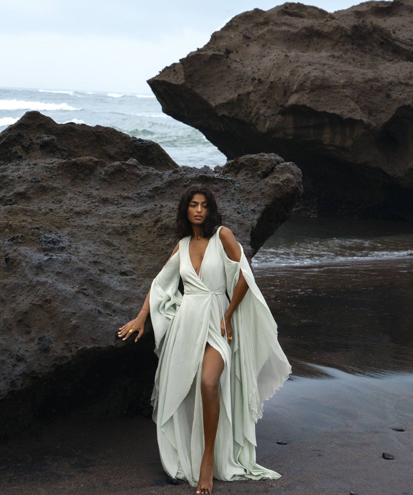 Model sitting in a tropical Bali garden wearing the ethereal ice blue Makosh goddess dress with mist blue silk wing sleeves. Textured organic crinkle cotton wrap gown for a celestial bridal look. Sustainable handcrafted wedding wear by AYA Sacred Wear.